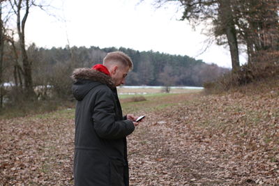 Side view of man using mobile phone while standing in forest against sky