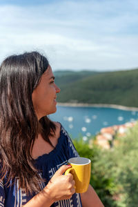 Portrait of happy young woman holding mug of coffee on balcony overlooking sea coast