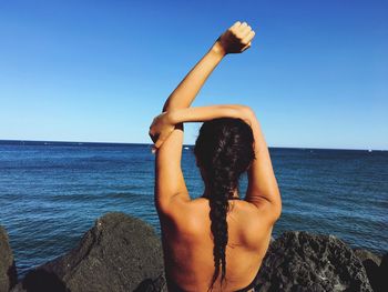 Rear view of young woman exercising on shore against clear blue sky