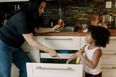 Mother pointing at daughter putting hand inside drawer in kitchen