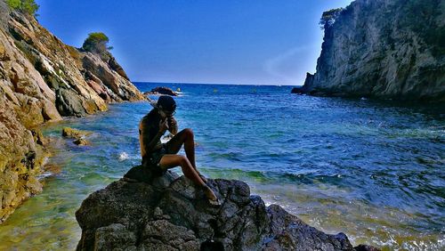 Man sitting on rock by sea against clear sky