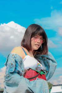 Low angle view of young woman holding purse while standing against blue sky