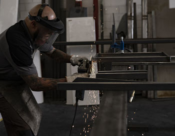 Side view of serious male welder using electric grinder and cutting metal detail in grungy workshop