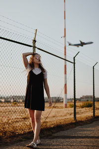 Full length of woman standing by chainlink fence against sky