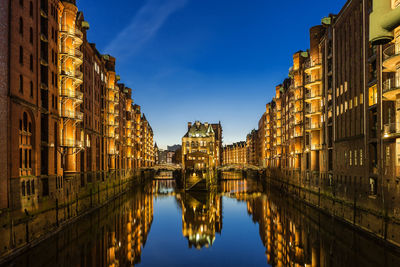 Canal amidst illuminated buildings against blue sky