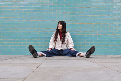 Young woman looking away while sitting with legs apart in front of brick wall