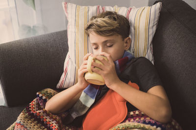 Ill boy with hot water bottle lying on sofa while drinking coffee