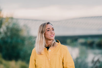 Smiling female in yellow raincoat standing in park and looking away while enjoying weekend