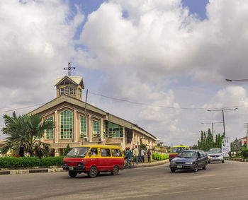 Cars on road by building against cloudy sky
