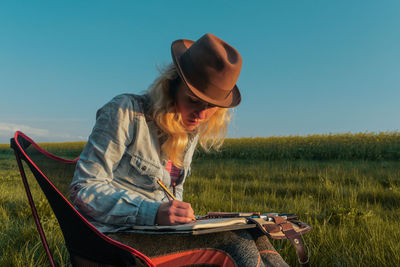 Woman drawing in book while sitting on field against clear sky