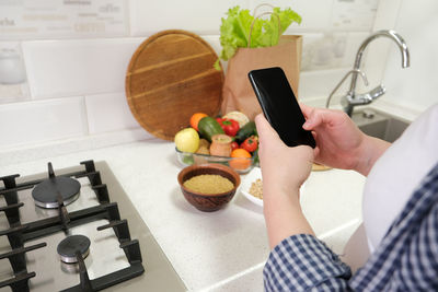 Female hand holding cell phone with empty screen on background kitchen table with grocery