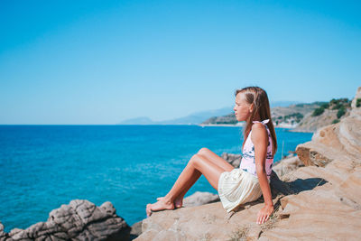 Woman sitting on rock by sea against clear blue sky