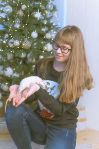 Girl holding rat while sitting against christmas tree