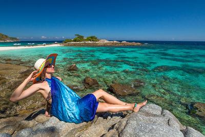 Woman sitting on rock by sea against blue sky