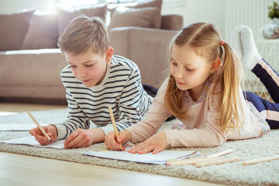 Siblings writing on paper while lying at home