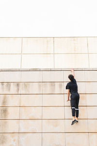 Back view male athlete in black clothes holding hand on stone border wall and hanging down while training on city street