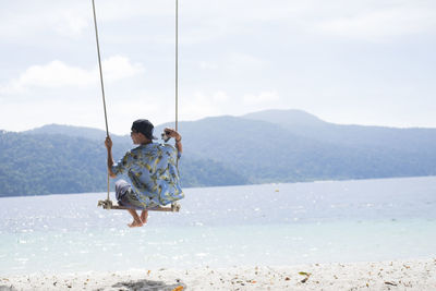 Rear view of mid adult man swinging at beach against sky