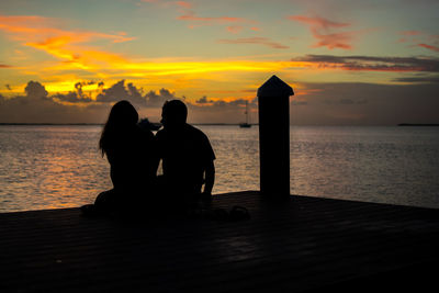 Silhouette couple sitting on pier in sea against orange sky during sunset