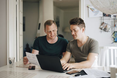 Smiling male homosexual couple paying bill through laptop on dining table at home