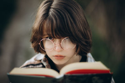 A teenage girl with short dark hair in glasses holds a book in her hands close to her face.