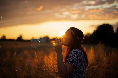 Young woman blowing bubbles on wheat field against cloudy sky during sunset