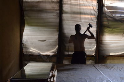 Rear view of shirtless man holding hammer while standing by window