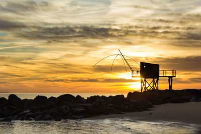 Silhouette fishing hut on beach against sky during sunset