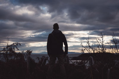 Rear view of man standing on field against cloudy sky at dusk