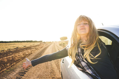 Young woman with arms outstretched leaning out of car window against clear sky