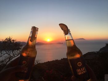 Close-up of hand holding beer bottle against sky during sunset