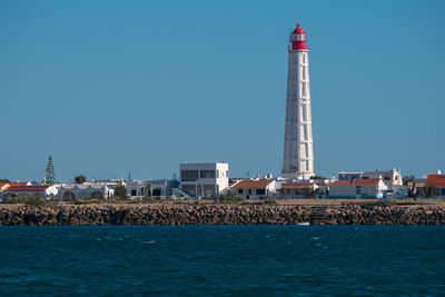 Lighthouse by sea against buildings against clear blue sky