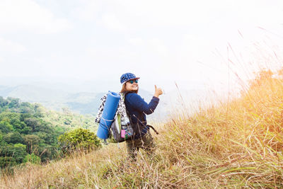 Rear view of woman with backpack showing thumbs up on field against clear sky