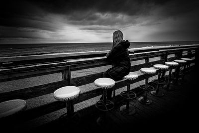 Side view of woman sitting at beach against cloudy sky