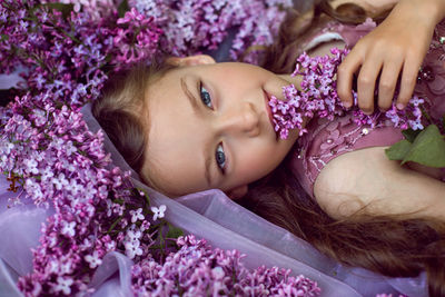 Child girl in a purple floral dress lies on the ground among lilacs on a veil in spring