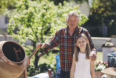 Portrait of confident grandfather and granddaughter standing by cement mixer in yard