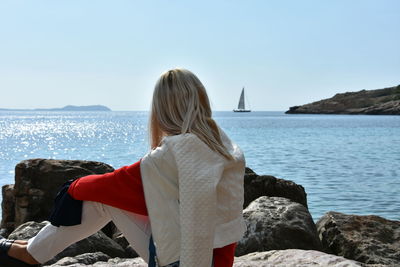 Side view of woman sitting on rocky shore against sky