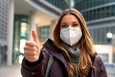 Woman wearing protective mask kn95 ffp2 showing thumb up with city background and looking at camera