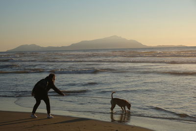 Full length of dog and girl on beach against sky during sunset