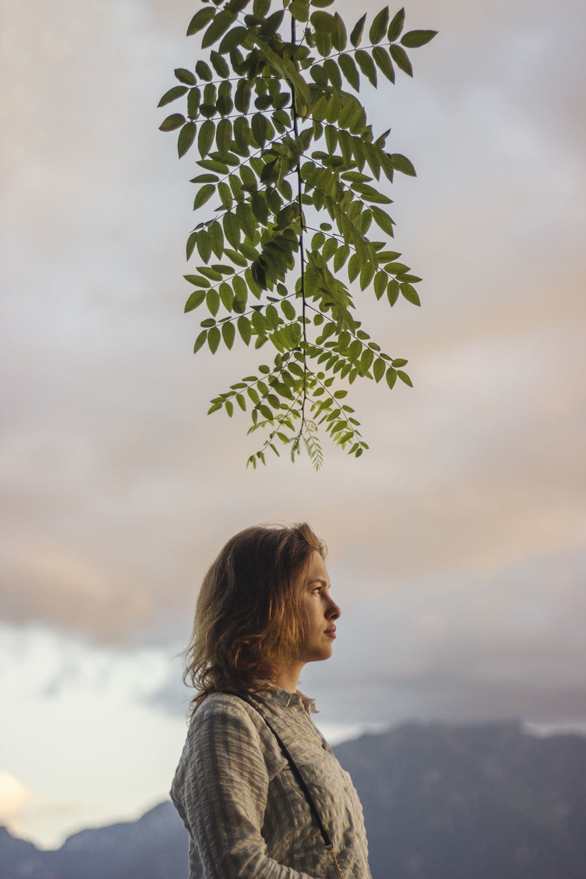 REAR VIEW OF WOMAN STANDING AGAINST TREE MOUNTAIN AGAINST SKY