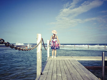 Rear view of girl standing by railing on jetty at sea against blue sky