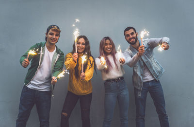 Portrait of cheerful friend holding sparkler sanding against wall