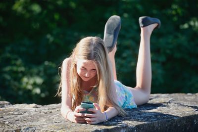 Girl using smart phone while lying on retaining wall outdoors
