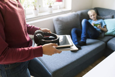 Midsection of teenager holding books and technology while brother sitting on sofa at home