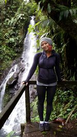 Full length portrait of woman standing against waterfall at forest