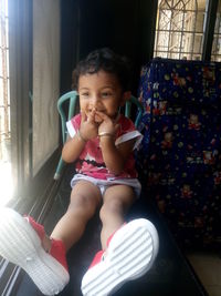 Cute happy girl with finger on lips sitting on window sill at home