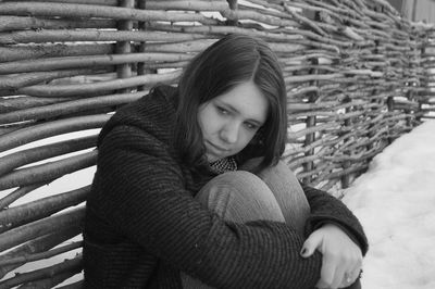 Thoughtful woman hugging knees while sitting against fence on snow covered field