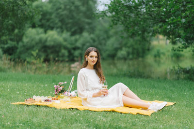 A beautiful young woman in a white dress drinks wine in the garden on a picnic. summer vacation