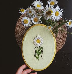 Close-up of hand holding embroidery art over white flowering plants