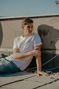 Young man looking away while sitting by wall outdoors