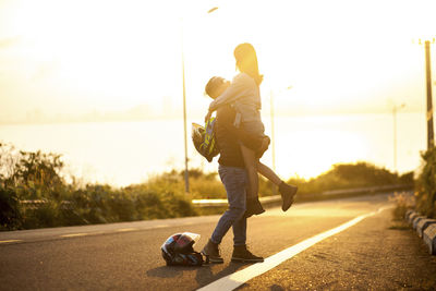 Man picking up woman while standing on road during sunset